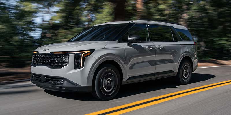 Gray 2026 Kia Carnival MPV Hybrid driving on a tree-lined road, shown in motion from a front-side angle.