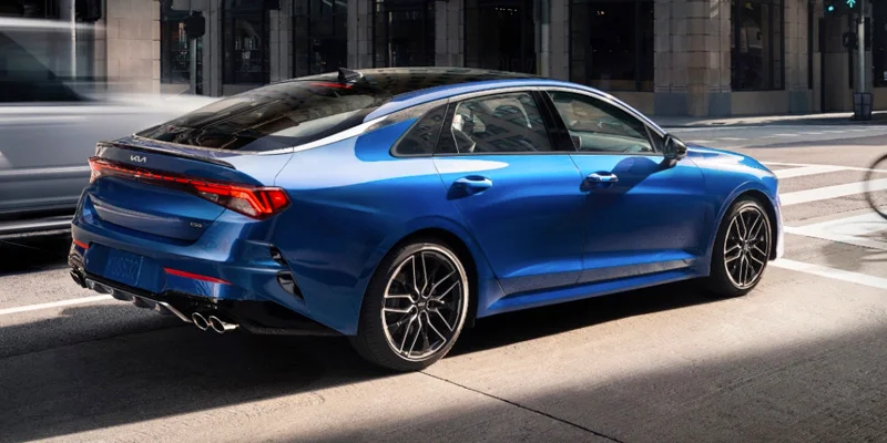 A blue Kia K5 driving through an urban street, showcasing its sleek rear design and sporty features. The vehicle is captured in motion, with blurred background to highlight its modern style.