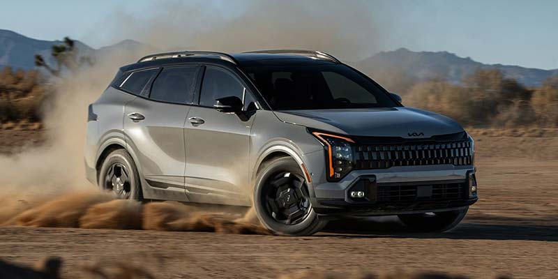 A silver Kia Sportage SUV driving through a dusty desert terrain, showcasing its dynamic handling and rugged design. The vehicle’s aggressive stance is emphasized as it kicks up dust on the road.