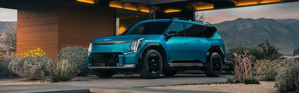 Teal 2026 Kia EV9 electric SUV parked in front of a modern home with mountain views at dusk.