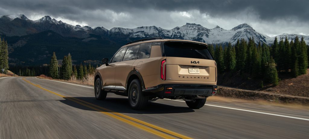 2026 Kia Telluride SUV driving on a scenic mountain highway with snow-capped peaks in the background.