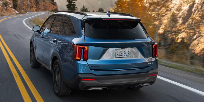 Blue 2026 Kia Sorento Hybrid SUV from the rear, driving along a scenic mountain highway with fall foliage.