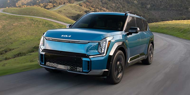 A blue Kia EV driving on a winding road surrounded by green hills.