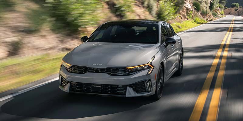 A sleek Kia Sedan driving on a winding road surrounded by trees.