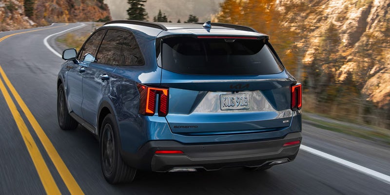 A blue Kia SUV driving on a winding road with mountains and autumn foliage in the background.