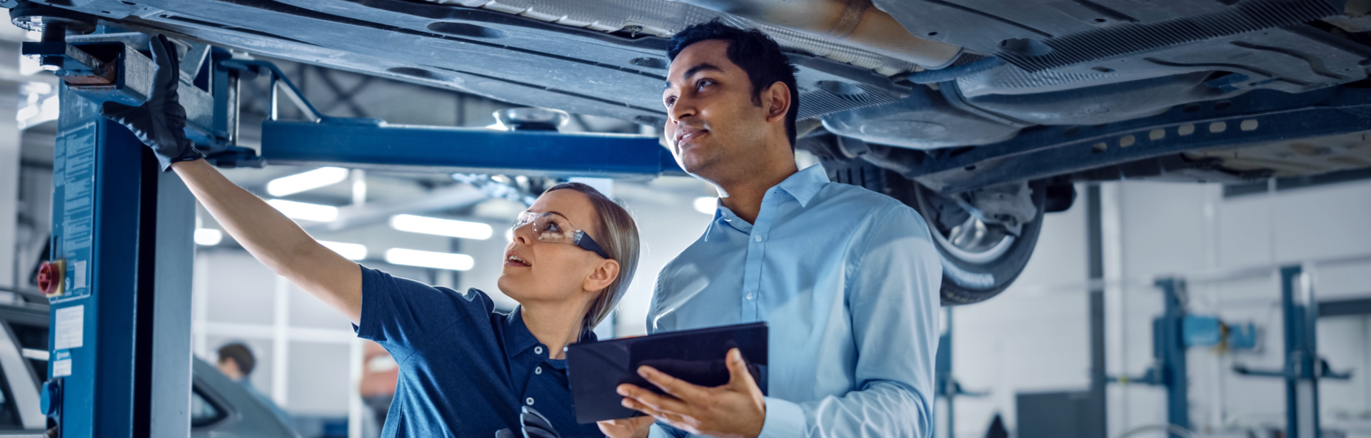Two auto mechanics inspecting a vehicle on a lift in a garage, one holding a tablet and the other pointing at an area under the car.