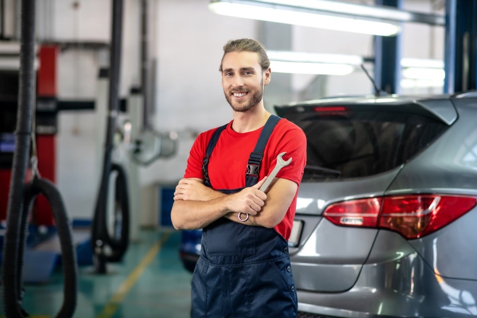 A mechanic smiling and standing with arms crossed in an auto repair shop, with a car in the background.